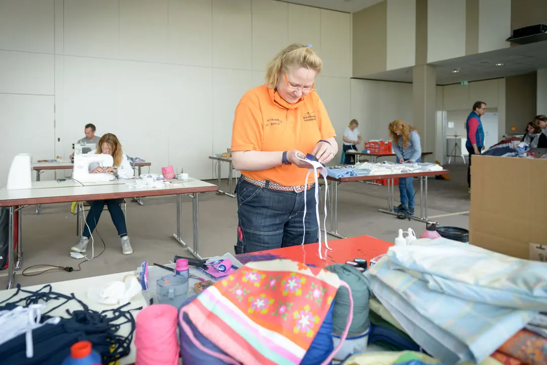 Eva Erkenberg hält Stoff in den Händen, im Hintergrund sind Teilnehmerinnen und Teilnehmer der Nähaktion fleißig