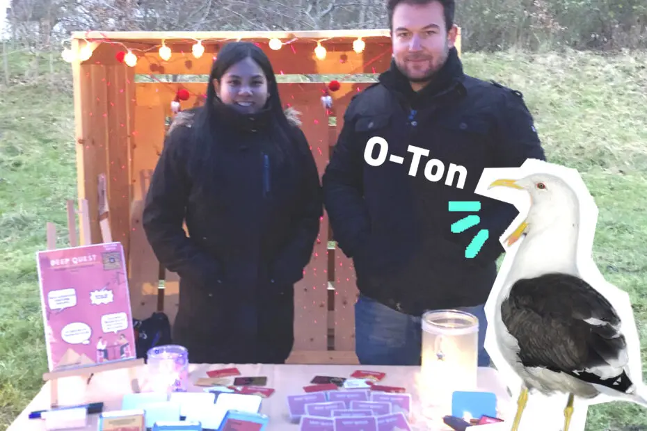 Link zu O-Ton: Deep Quest. Eine Frau und ein Mann aus dem Team Deep Quest stehen an einem Tisch mit ihrem selbst entwickelten Kartenspiel