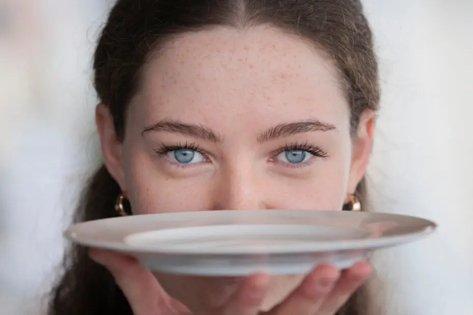 Junge Frau hält einen Teller vor ihr Gesicht und blickt buchstäblich über den Tellerrand
