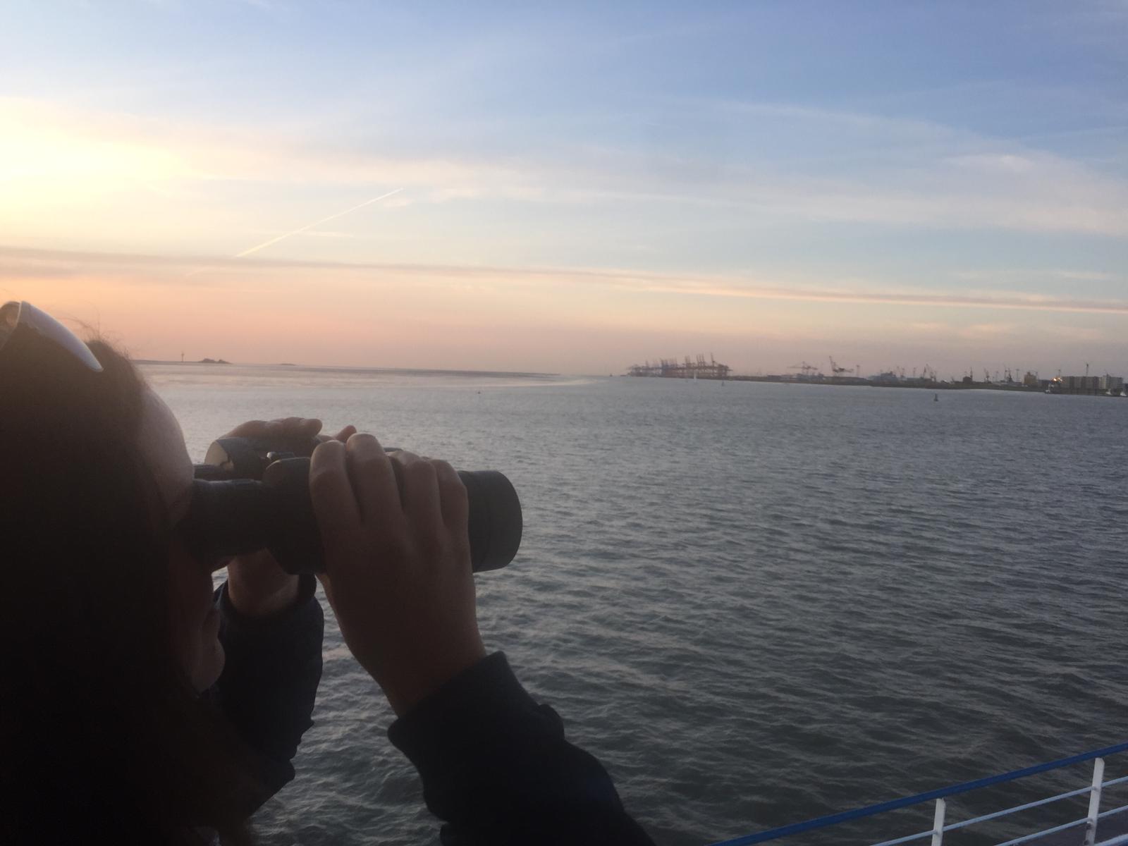 Lisa Keller hält ein Fernglas und blick auf über Wasser auf den Hafen