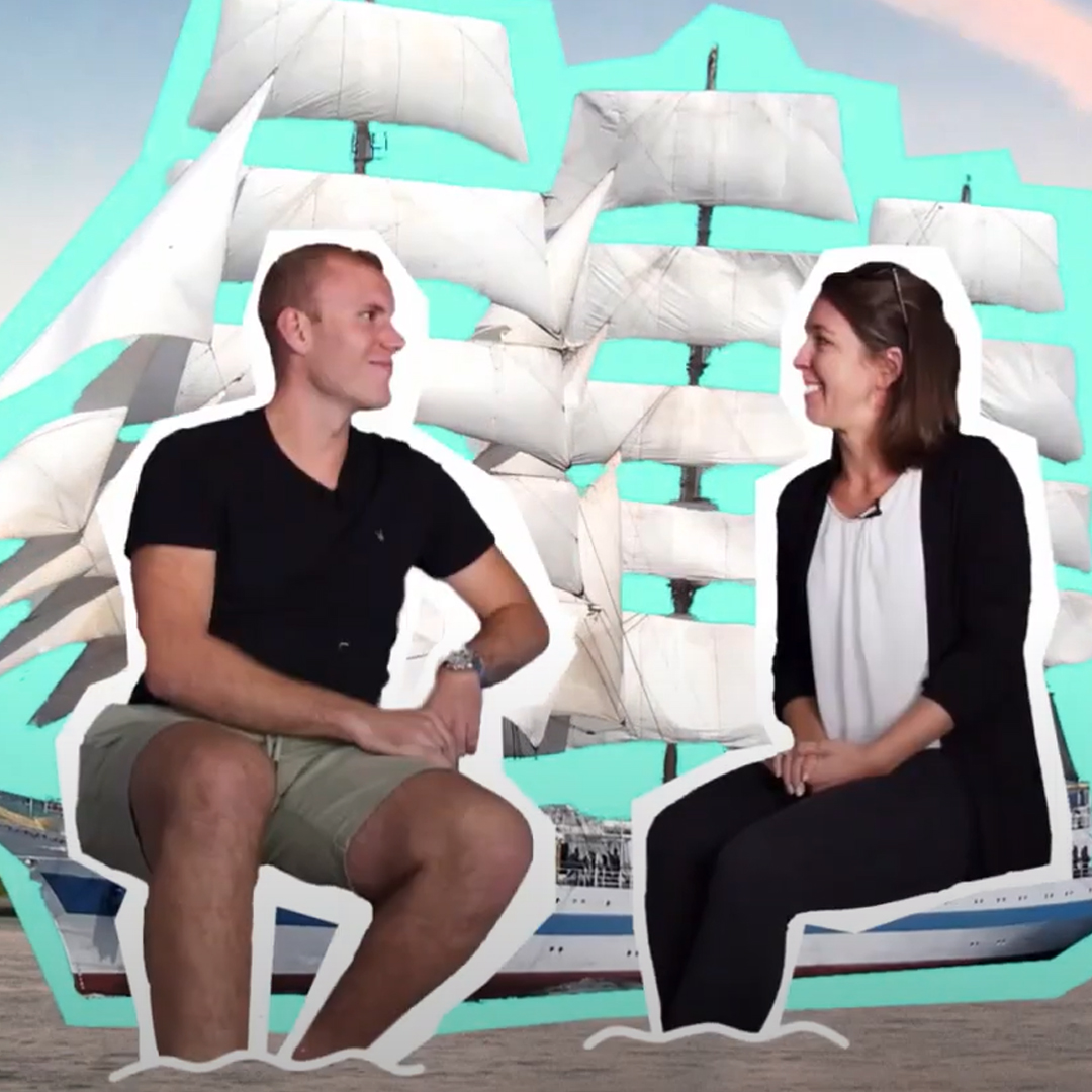 Eine Frau und ein Mann sitzen sich im Gespräch gegenüber, im Hintergrund die Montage eines Segelschiffs mit weißen Segeln