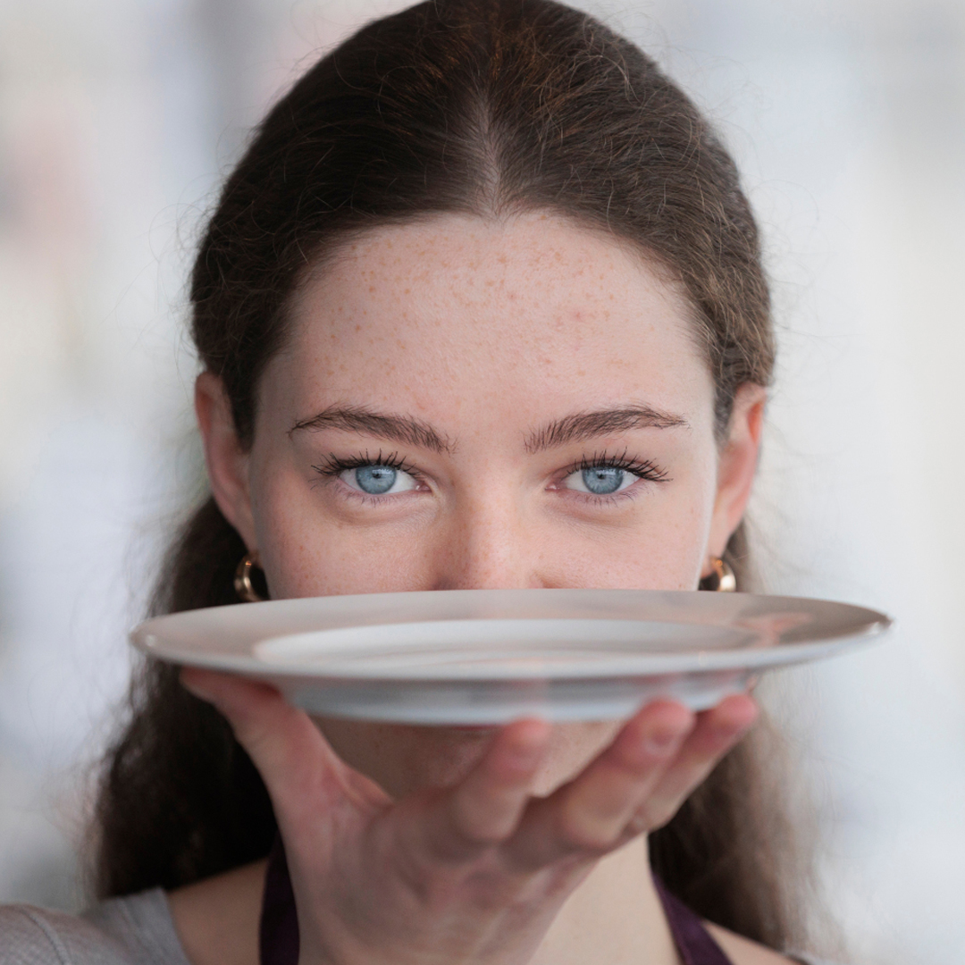 Junge Frau hält einen Teller vor ihr Gesicht und blickt buchstäblich über den Tellerrand